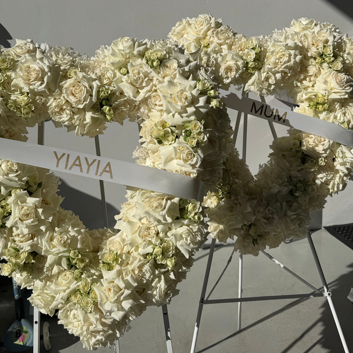 Three large heart-shaped floral wreaths on white metal stands, decorated with cream and white roses and green accents, with personalized names 'YIAYIA', 'MUM', and another name on white ribbons.