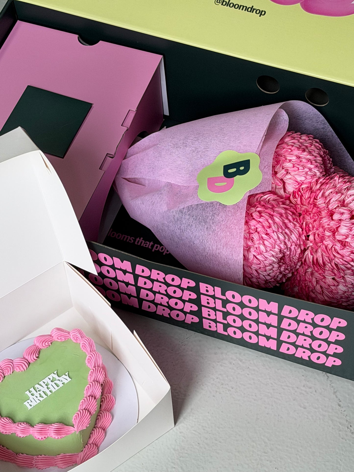 Pink and green heart-shaped cake with 'Happy Mother's Day' text, pink socks, and 'Bloom Drop' branding.