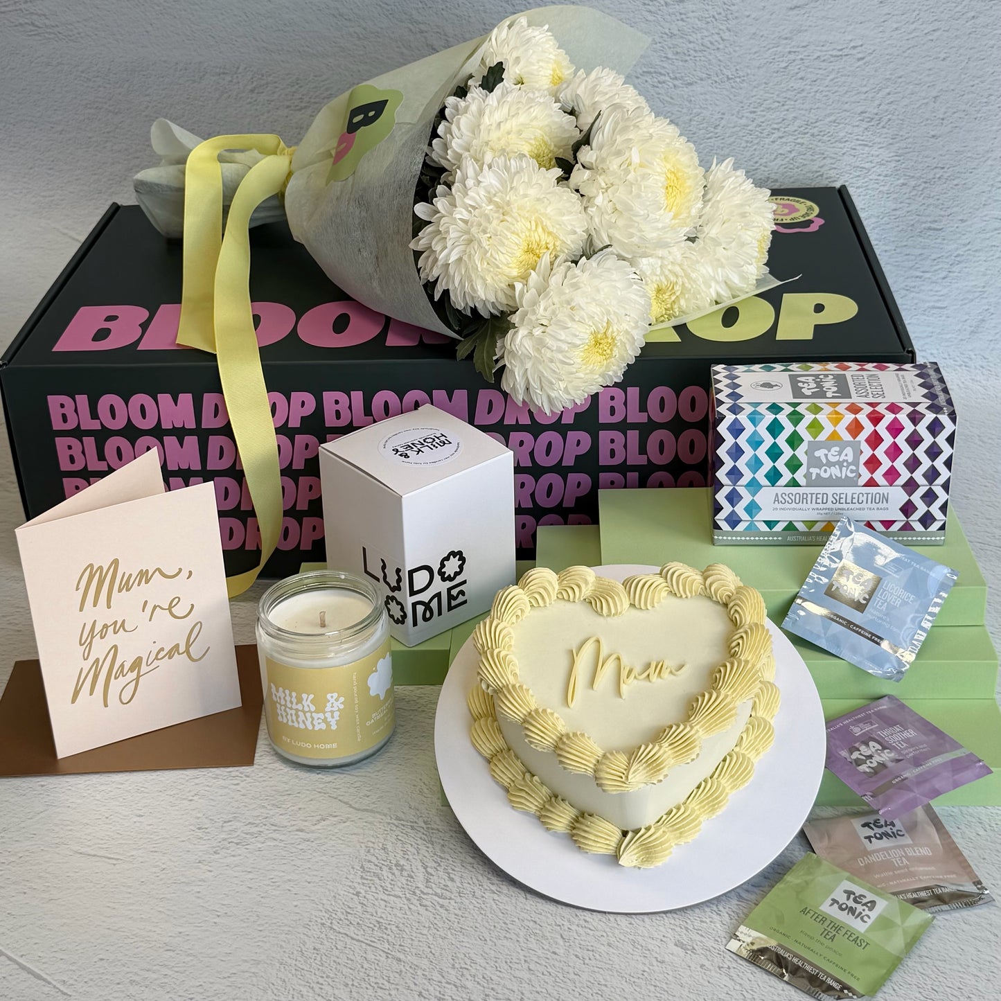 Heart-shaped cake, bouquet of flowers, candle, and cards on a table with a Bloom & Pop box in the background.