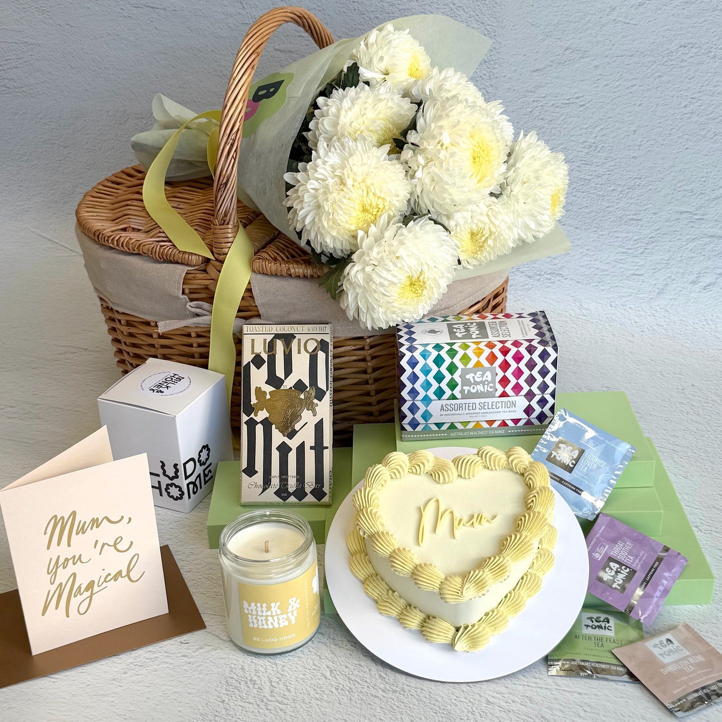 Heart-shaped cake with 'Mum' on a white surface, surrounded by flowers, cards, and candles.