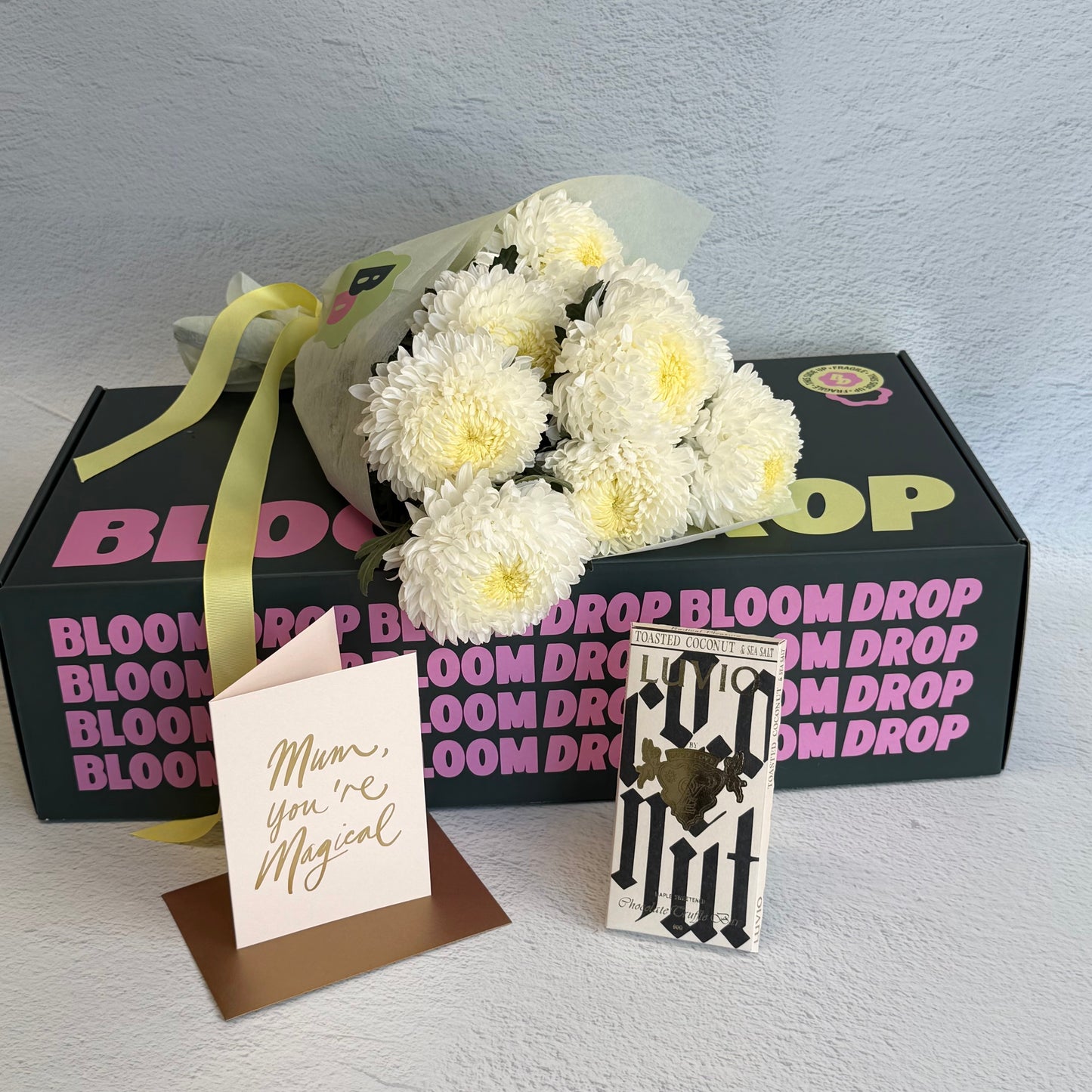 Bouquet of white flowers with a card and decorative item in front of a Bloom Drop box.